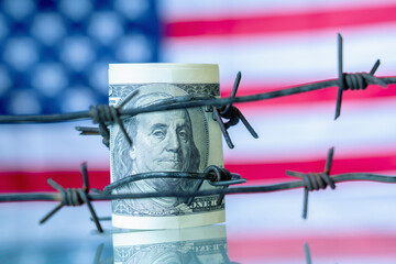 US Dollar currency wrapped in barbed wire against flag of United States as symbol of economic...