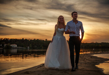 Young married couple walk together on a sandy beach, on background of dramatic sunset sky over green forest