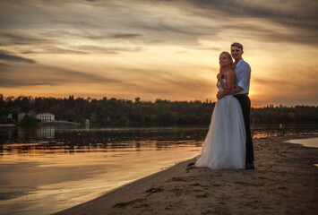 Young married couple walk together on a sandy beach, on background of dramatic sunset sky over green forest