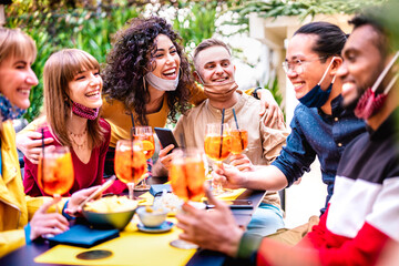 Friends toasting drinks at cocktail bar with face mask - New normal life concept with happy people having fun together drinking spritz at restaurant - Bright end vivid filter - Shallow depth of field