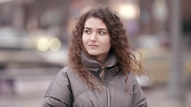 Portrait of a young woman with curly hair, posing on a city street on a cold day against the background of evening lights and cars.