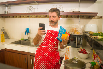 home cook at work ! young happy and attractive home cook man in red apron following online recipe on internet mobile phone cooking healthy food relaxed at nice cozy kitchen