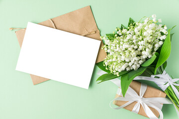 Blank greeting card with spring lily of the valley flowers bouquet and gift box on pastel green background. Flat lay