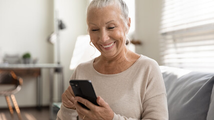 Close up overjoyed mature woman using phone, sitting on couch, browsing smartphone apps, looking at...