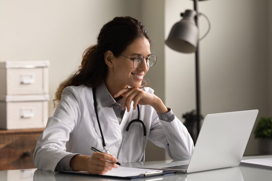 Smiling professional female doctor wearing glasses and uniform taking notes in medical journal, filling documents, patient illness history, looking at laptop screen, student watching webinar