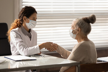 Side view smiling female doctor wearing face mask and mature patient shaking hands at meeting in office, senior woman making health insurance deal, successful medical checkup results, healthcare