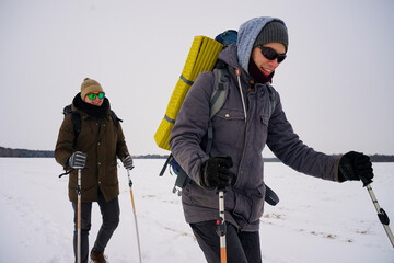 Fototapeta na wymiar Two guys walk through loose snow during a winter expedition. They carry large backpacks, warm jackets. They hold trekking sticks in their hands.