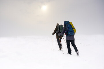 Two guys walk through loose snow during a winter expedition. They carry large backpacks, warm jackets. They hold trekking sticks in their hands.