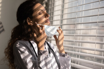 Close up tired female doctor taking off medical face mask, standing near window in hospital office after long workday or surgery, breathing fresh air deep, physician holding face protection