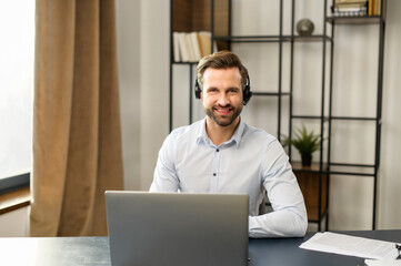 Smiling young bearded man wearing headset looking at camera, positive hipster in formal wear working in the customer service department, making and receiving calls, sitting in the home office