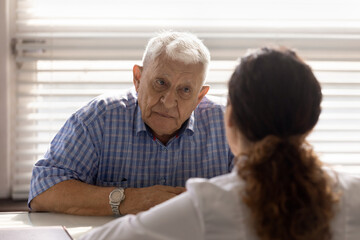 Close up anxious serious old man listening to female doctor at meeting in hospital, therapist...