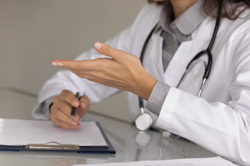 Close up hands of female doctor wearing uniform with stethoscope consulting patient, filling medical document, patient card on clipboard, explaining treatment, prescriptions, giving recommendations