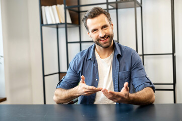 Young bearded hipster guy in casual clothes sitting at desk, looking at camera, moving hands,...
