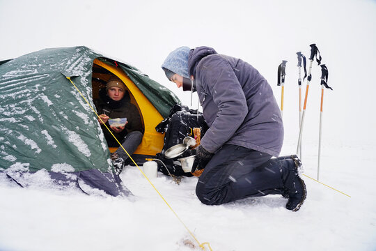 Guys Near The Tent In A Snowy Brownstone Are Preparing Food During A Winter Trip.