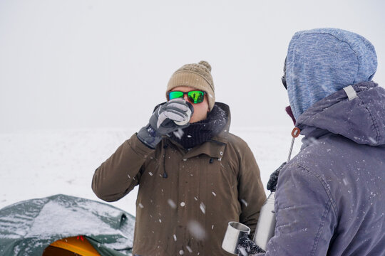 The guys drink hot tea from thermo mugs and pour water from a thermos. They stand by the tent during the winter expedition.