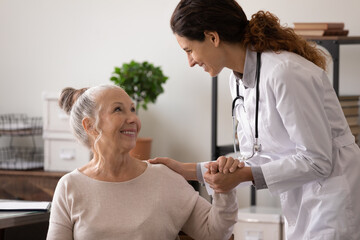 Smiling mature patient and caring female doctor holding hands, discussing good news, successful treatment, caregiver wearing uniform supporting senior woman in hospital, psychological help concept