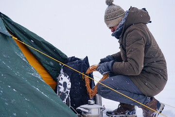 A tourist sits near a tent set on the snow, he takes out a Cord from a backpack for additional fixation of the tent during a winter storm