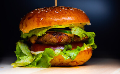 juicy tasty hamburger with bread loaf, meat cutlet, cheese and vegetables with herbs on a wooden board on a black background