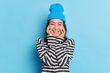 Portait of pleasant looking cheerful Asian woman keeps hands on cheeks smiles gently wears striped jumper stylish hat expresses sincere emotions isolated on blue background. Positive face expressions