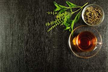 Tulsi or holy basil tea in transparent glass cup with fresh tulsi leaf and dried leaf in wooden bowl on black wooden background top view. Ayurvedic medicine in India.