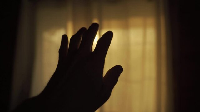 Hand moves slowly in dark room with warm sun shining through window curtains. Slow motion movement of a hand with slight flare. Sun entering the house in early morning, sunrise or sunset time of day