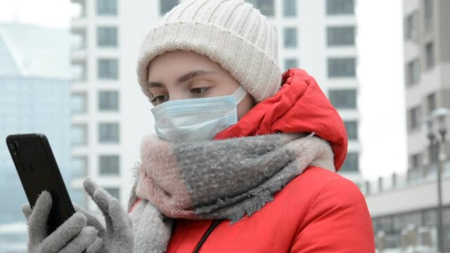 Hipster girl in a protective mask with a phone in her hands writes a message outside