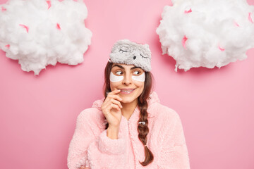 Pretty satisfied European girl smiles gently has curious face expression enjoys comfort and serene atmosphere at home wears sleepmask with pajama isolated over pink background going to sleep