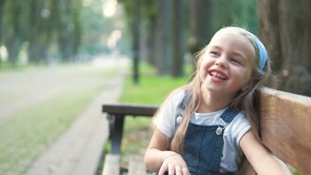 Little happy child girl sitting on a bench smiling happily in summer park.