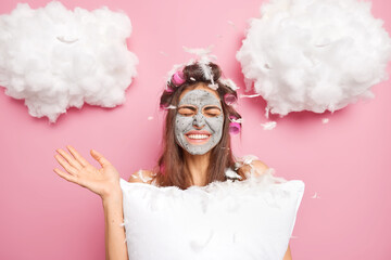 Positive woman has joyful mood raises palm closes eyes poses indoor with lot of feathers around after pillow fisht makes hairstyle with rollers applies clay mask on face. Sleeping wake up beauty