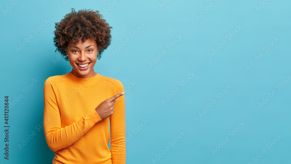 Sticker Smiling black curly haired woman in casual jumper points aside shows space for your advertisement or promo promots big sale has special offer isolated over blue background. Great suggestion.