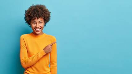 Smiling black curly haired woman in casual jumper points aside shows space for your advertisement or promo promots big sale has special offer isolated over blue background. Great suggestion. - obrazy, fototapety, plakaty