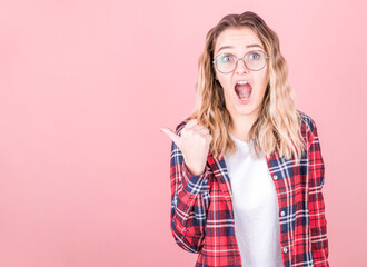 Happy excited woman with wavy blond hair in plaid shirt pointing at copy space with finger isolated on pink background