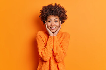 Fototapeta na wymiar Portrait of pleasant looking dark skinned woman keeps hands on face admires something smiles gently and looks directly at camera wears sweater isolated over vivid orange background. Monochrome