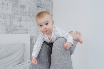 Cute little caucasian baby lying on legs of dad, looking at camera on bed, modern white and gray interior at home. Leisure activities with parent. Copy space.