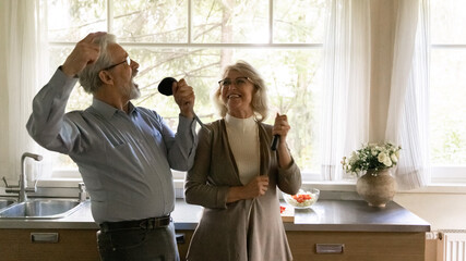 Excited happy mature family couple singing at utensils and having fun while cooking dinner...