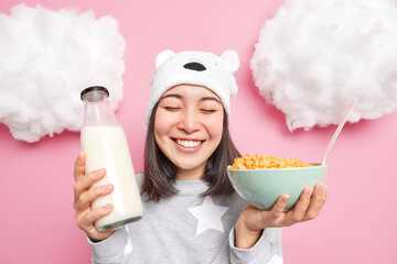 Positive Asian girl enjoys good morning has breakfast eats cereals with milk wears pajama and soft...