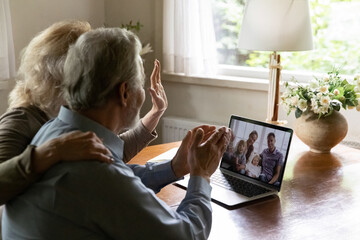 Mature pensioner grandparents couple talks to children and grandchildren, elder OAP use laptop for video call, wave hello. Screen view of happy young 60s parents and two kids during covid 19 concept - obrazy, fototapety, plakaty