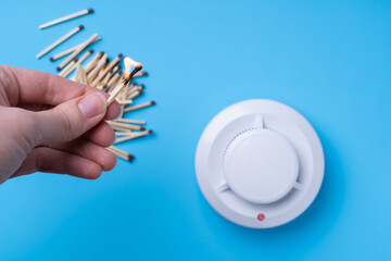 Hand with a burning match and a fire detector on a blue background. Fire safety concept.