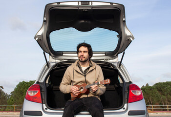 Fototapeta na wymiar Smiled young man with ukulele in his hands sitting in the trunk. Young man playing ukulele
