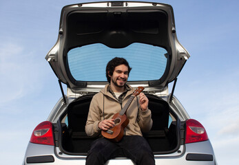 Smiled young man with ukulele in his hands sitting in the trunk. Young man playing ukulele