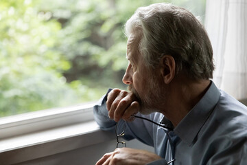 Pensive mature 70s aged man in deep thoughts holds glasses, looks out window. Thoughtful older...