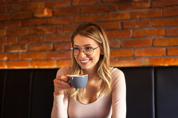 Beautiful young blonde girl sitting in a restaurant drinking coffee, listening carefully while wearing glasses and smiling in front of a brick wall