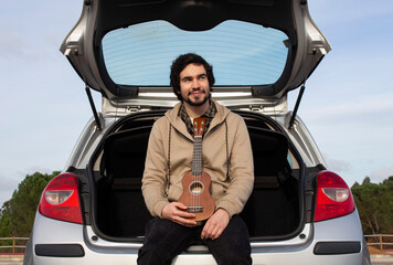 Smiled young man with ukulele in his hands sitting in the trunk. Young man playing ukulele