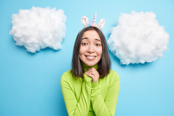 Hopeful beautiful young Asian woman keeps hands in pray looks with mercy expression asks for help has eyes full of hope wears casual green jumper isolated over blue background with clouds above