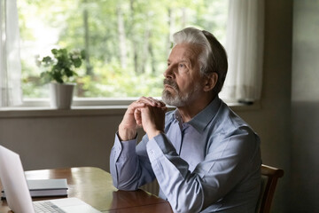Pensive mature grey haired 60s - 70s aged man sitting at table with laptop at home, looking away...