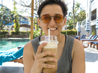 Young woman with short hair, sunglasses is sitting and drinking iced coffee by the pool.