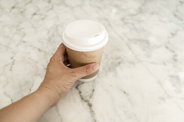 Woman hand is holding paper cup of hot coffee on white marble table.