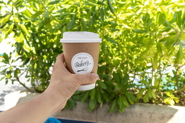 Woman hand holding delivered paper cup of hot coffee on green plants background.