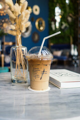Iced coffee with straw on the white marble table with book and glass vase of dried decoration plant.
