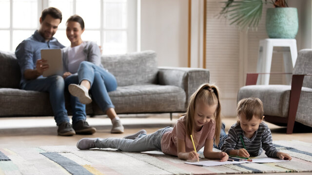 Happy young couple and preschooler children enjoying weekend at home. Kids rest on heating floor, playing and drawing, parents relaxing on couch, using tablet. Family leisure time in modern cozy house